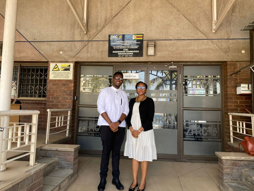 Two people, a man in a white shirt and black pants and a woman in a white dress and black jacket, stand smiling in front of the entrance to the ZHRC offices on a sunny day.