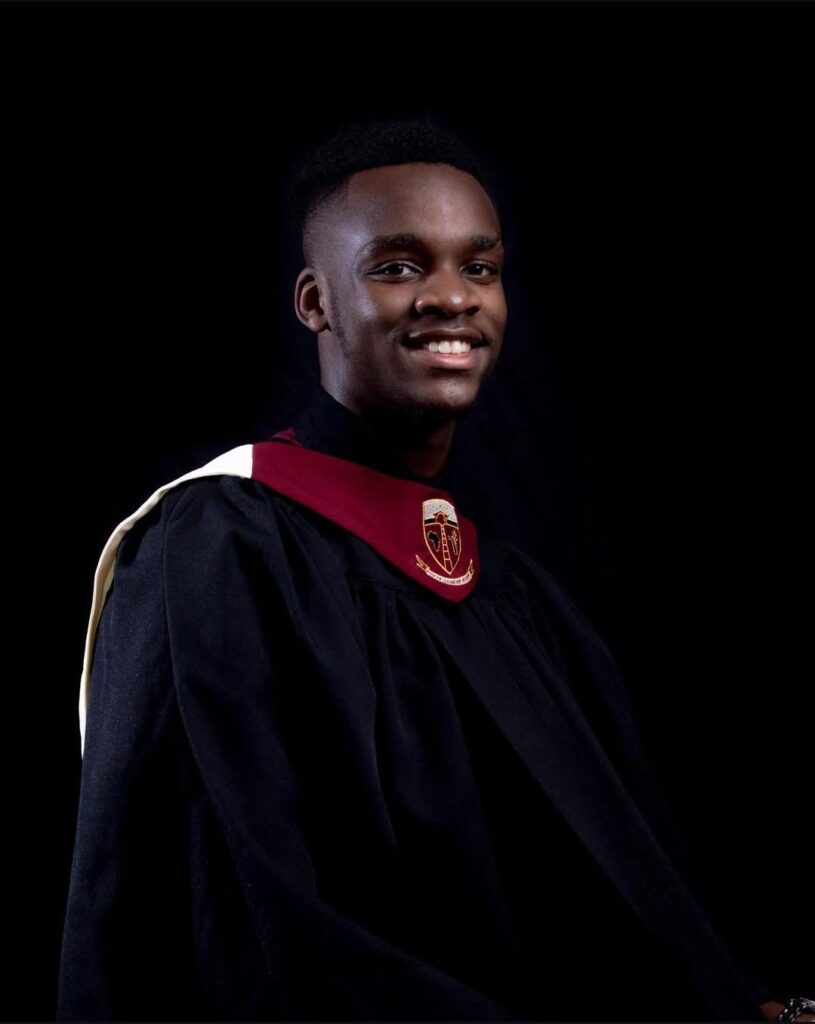 A young man in graduation attire with a maroon and gold stole sits and smiles against a black background.